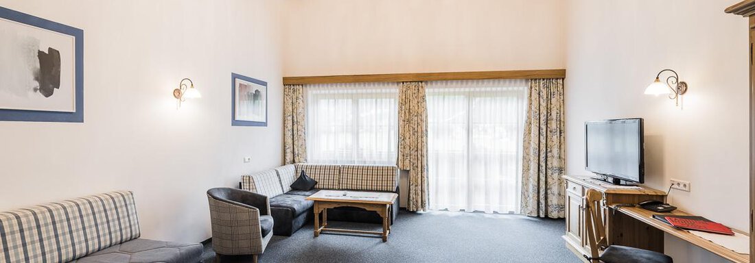A spacious living room with high ceilings and wooden beams. There is comfortable seating, a television, and large windows with curtains. - Andre Arnold - Boutique Hotel - Sölden
