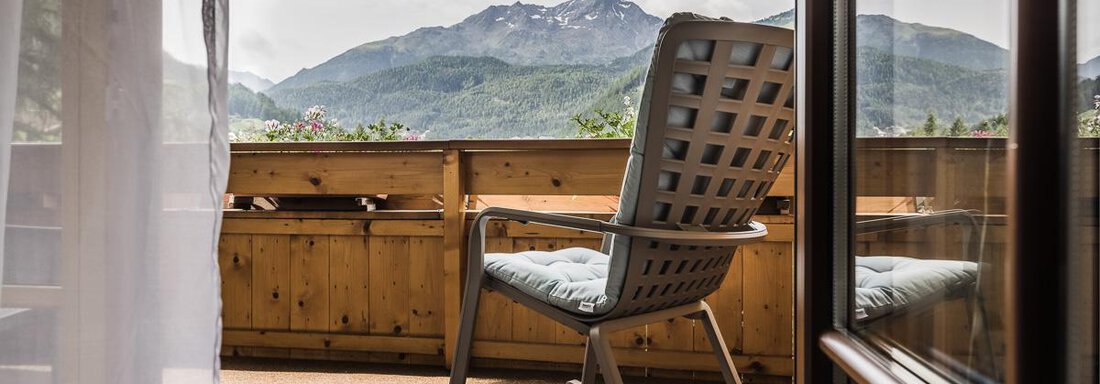 A rocking chair on a balcony overlooking the mountains. The surroundings are green, and the clouds give the scene a tranquil atmosphere. - Andre Arnold - Boutique Hotel - Sölden