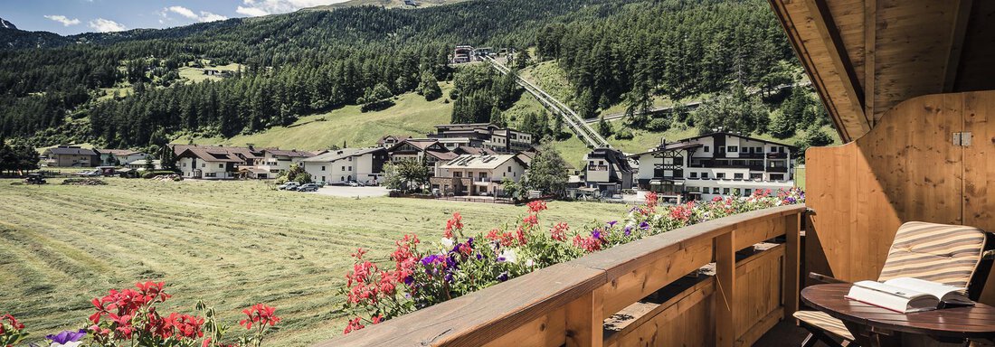 A terrace with a wooden railing and cozy chairs, surrounded by colorful flowers. In the background, a picturesque mountain landscape stretches out with green meadows and houses. - Andre Arnold - Boutique Hotel - Sölden