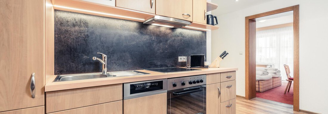 A modern kitchen with wooden cabinets and stainless steel appliances. In the background, a pleasant living area is visible. - Andre Arnold - Boutique Hotel - Sölden
