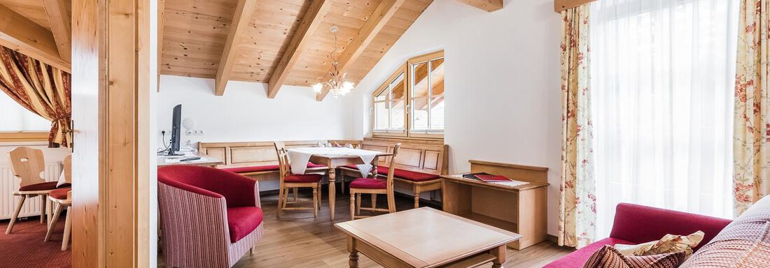 A cozy living space with a wooden beam ceiling, red sofas, and a dining table. Large windows let in plenty of light and create an inviting atmosphere. - Andre Arnold - Boutique Hotel - Sölden