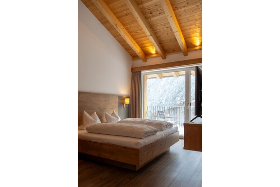 A modern bedroom with a wooden ceiling and a large window. The room is bright and cozily furnished, with a comfortable bed and a television. - Anraitl Appartements Sölden - Sölden
