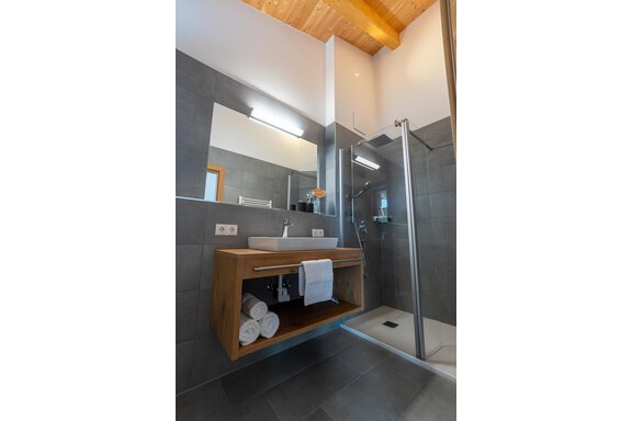 A modern bathroom with grey tiles and a glass shower. The sink area is equipped with wooden design and fresh towels. - Anraitl Appartements Sölden - Sölden