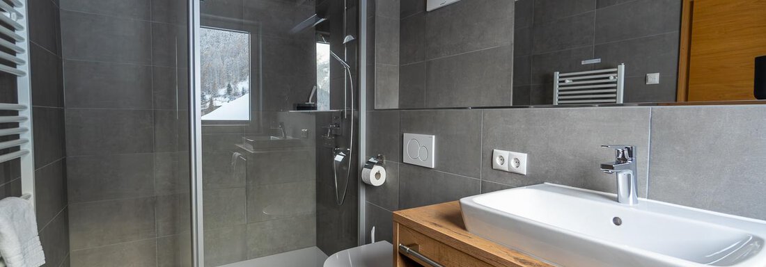 A modern bathroom with elegant gray tiles and a shower. The hand wash basin made of white ceramic material sits on a wooden dresser. - Anraitl Appartements Sölden - Sölden
