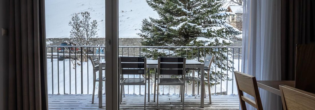 A cozy balcony with a table and chairs, from which one overlooks a snowy landscape. In the background, a green tree and snow-covered hills can be seen. - Anraitl Appartements Sölden - Sölden