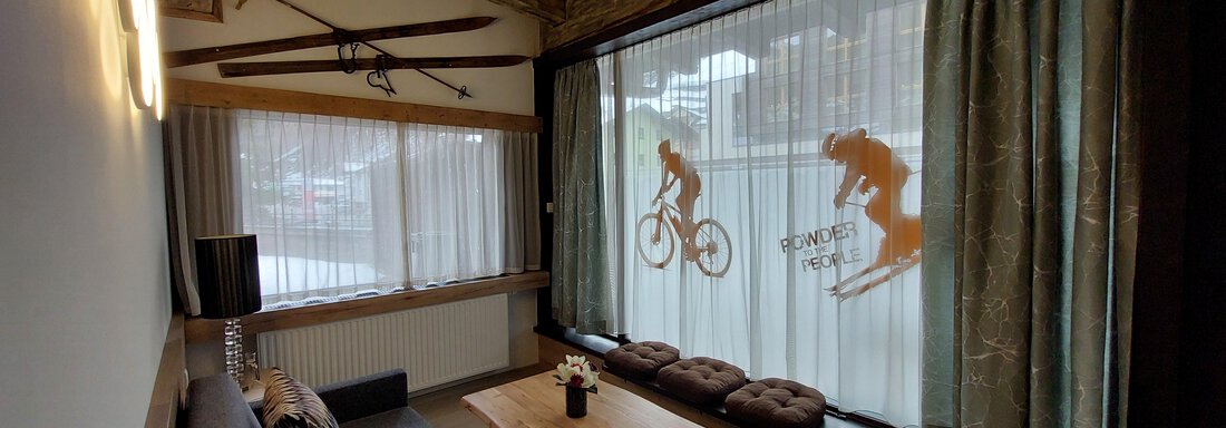 Wohnen | © Apart 77 (CC C) A cozy living space with a wooden ceiling and large windows. The windows feature silhouettes of cyclists and skiers. - Apart 77 - Sölden