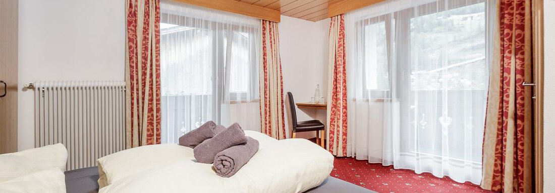A cozy room with a large bed and decorative pillows. There are windows with curtains and a small desk. - Apart Garni Enzian - Sölden