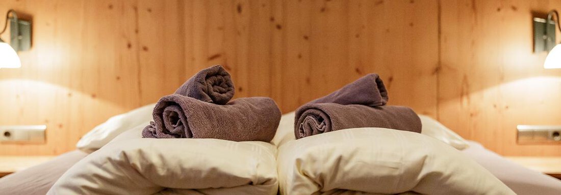 A cozy bedroom with stacked pillows and towels. The wood paneling and soft lighting create a warm atmosphere. - Apart Garni Enzian - Sölden