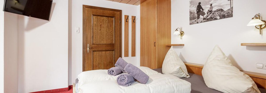 A cozy hotel room with wooden walls and a large window. There is a double bed with pillows and fresh towels, as well as a television on the wall. - Apart Garni Enzian - Sölden