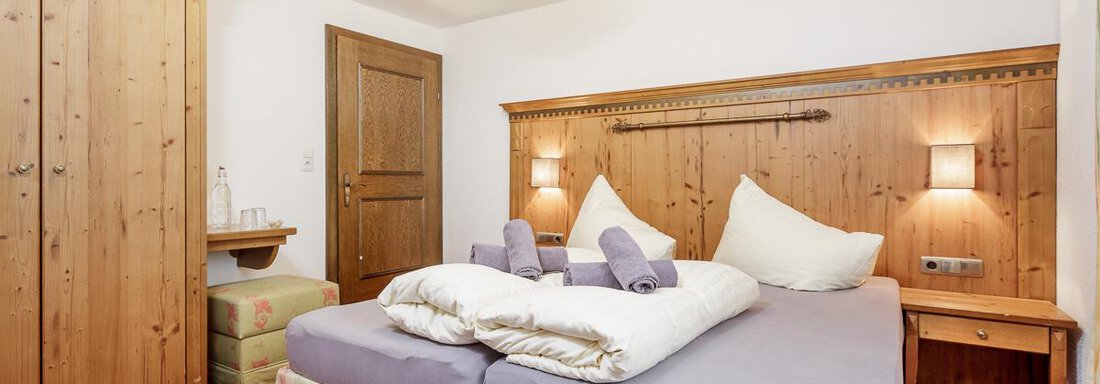 A cozy bedroom with a large double bed and wood-paneled walls. The room is simply and invitingly furnished, with a wardrobe and a small desk. - Apart Garni Enzian - Sölden