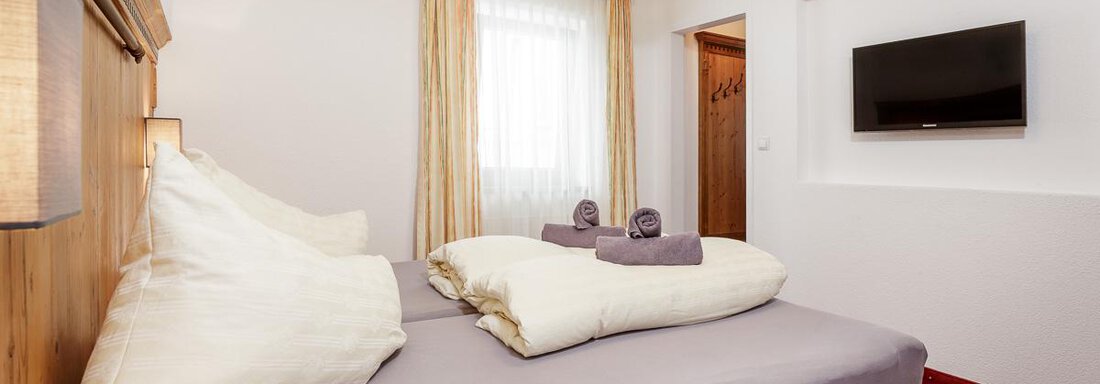 A cozy bedroom with a large double bed and soft pillows. A television hangs on the wall and there is plenty of daylight coming through the window. - Apart Garni Enzian - Sölden