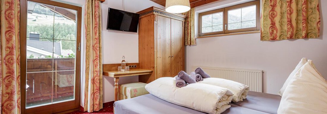 A cozy bedroom with wooden ceilings and carpet flooring. The furnishings include a double bed, a table, and a balcony with a view. - Apart Garni Enzian - Sölden