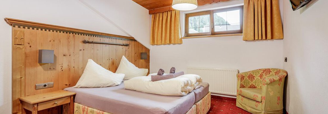 A cozy bedroom with a wooden bed, decorative pillows, and a comfortable armchair. The room has a warm atmosphere with wood paneling and red carpet flooring. - Apart Garni Enzian - Sölden