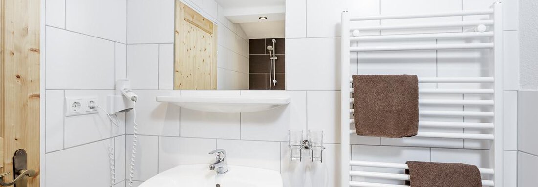 A modern bathroom with white tiles and a round sink. Towels hang on a radiator, and a large mirror reflects the room. - Apart Garni Enzian - Sölden