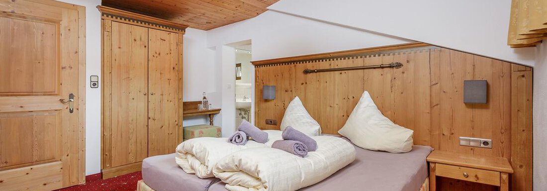 A cozy bedroom with a double bed and wooden furniture. The floor is red carpet and the walls are light, with a warm light from the lamp. - Apart Garni Enzian - Sölden