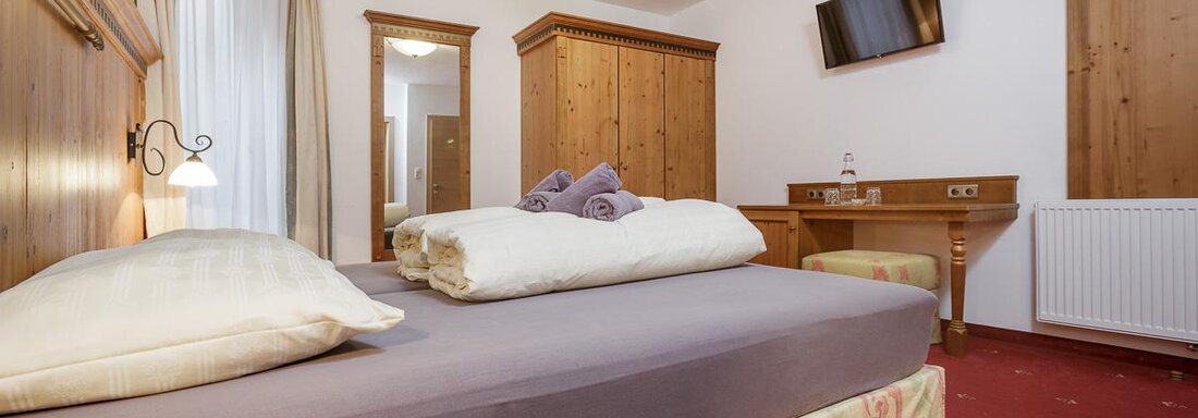 A cozy hotel room with a large bed and wood details. The room is bright and well-furnished, with a television and a desk. - Apart Garni Enzian - Sölden