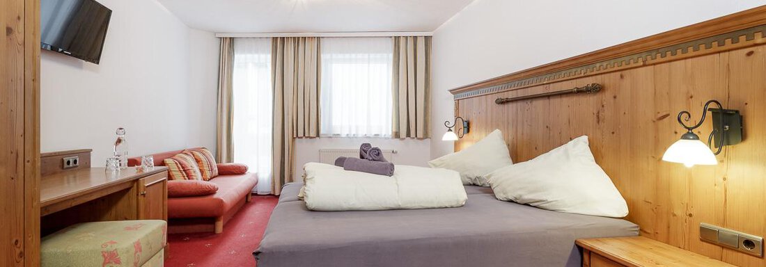 A cozy hotel room with a large bed and wooden paneling. There is also a television, a small table, and a sofa. - Apart Garni Enzian - Sölden