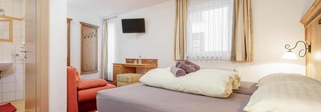 A cozy hotel room with a double bed, an armchair, and a television. The room has light curtains and a warm carpet. - Apart Garni Enzian - Sölden