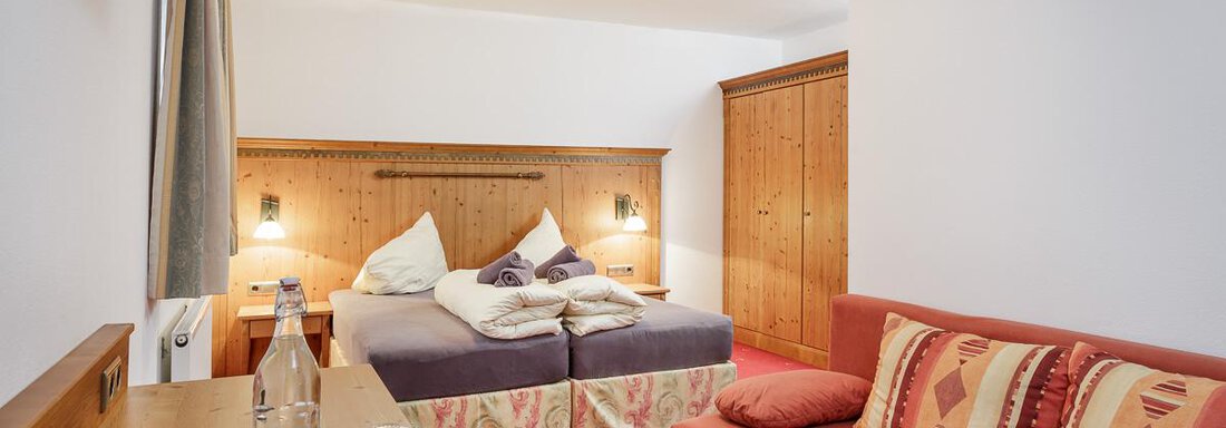 A cozy hotel room with a double bed and a red couch. The room is bright and inviting, with wooden furniture and a small table. - Apart Garni Enzian - Sölden