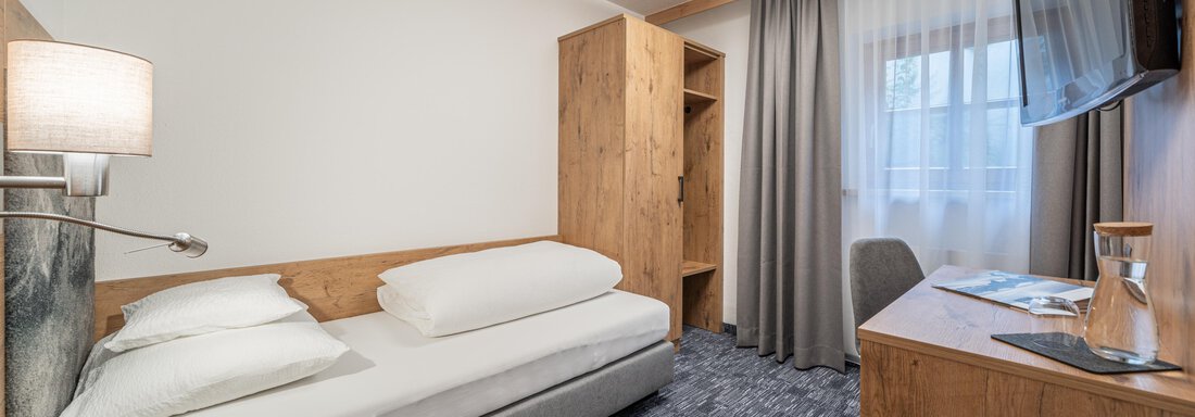 2 Einzelzimmer mit gemeinsamen Bad/Wc A modern hotel room with a single bed and wooden details. The windows are covered with curtains, and there is a television as well as a small desk. - Apart Hotel Garni Alt Kaisers - Sölden