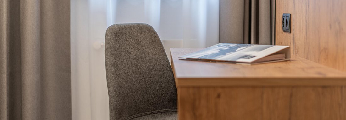 A modern desk with a gray chair and an open magazine. Bright curtains hang in front of the window. - Apart Hotel Garni Alt Kaisers - Sölden