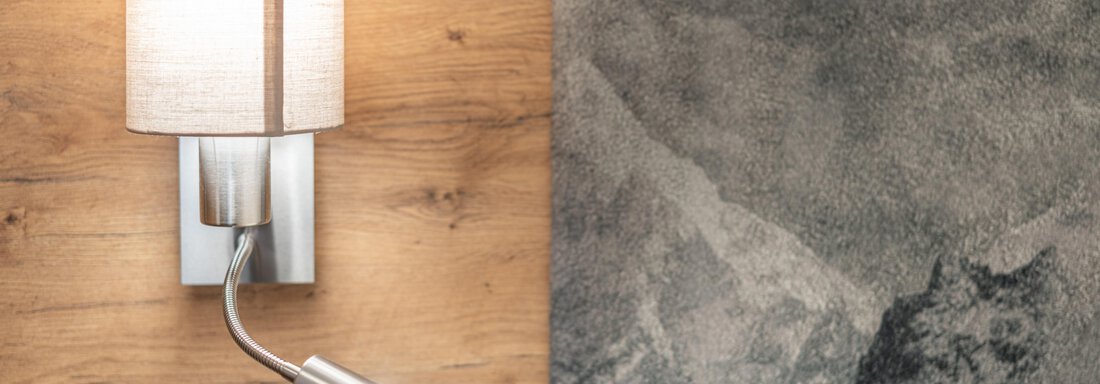 A modern wall lamp with an elegant shade on a wooden wall. Next to it hangs a painting with mountain motifs in shades of gray. - Apart Hotel Garni Alt Kaisers - Sölden