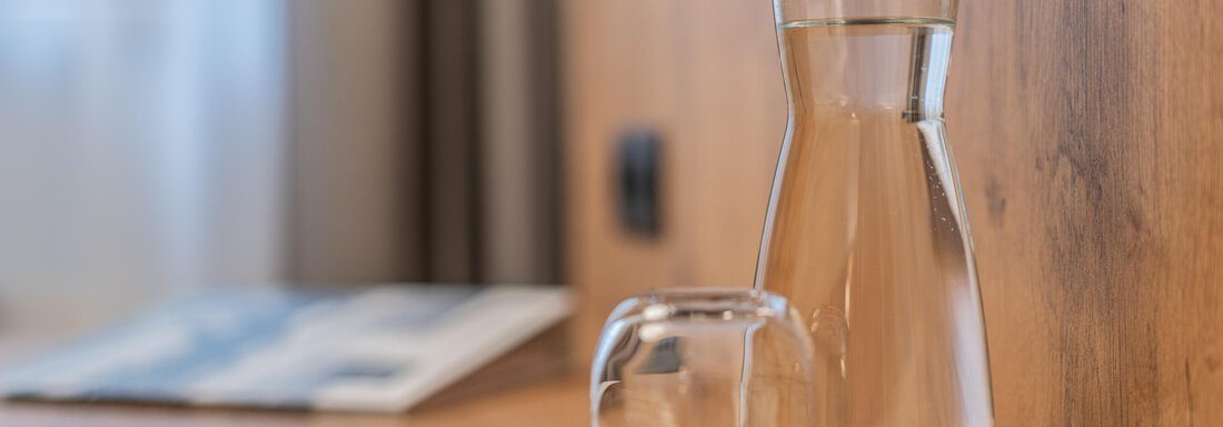 An elegant glass carafe with a cork lid stands on a wooden table. Next to it, there is a small glass on a black coaster. - Apart Hotel Garni Alt Kaisers - Sölden