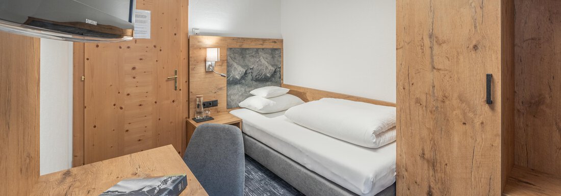 A modern hotel room with wooden decor and a comfortable single bed. There is a desk and a television for added comfort. - Apart Hotel Garni Alt Kaisers - Sölden
