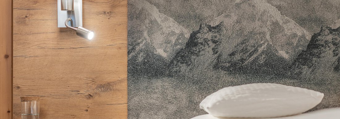 A cozy bedroom with a wooden panel and a mountain motif above the bed. Next to the bed is a modern wall lamp and a glass carafe. - Apart Hotel Garni Alt Kaisers - Sölden