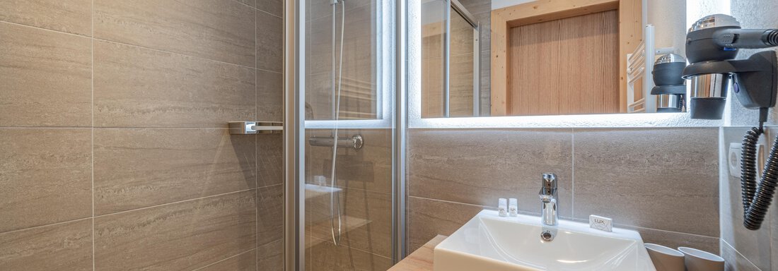 A modern bathroom with a shower and a sink made of white ceramic. The walls are in neutral tones, and there are wooden details. - Apart Hotel Garni Alt Kaisers - Sölden