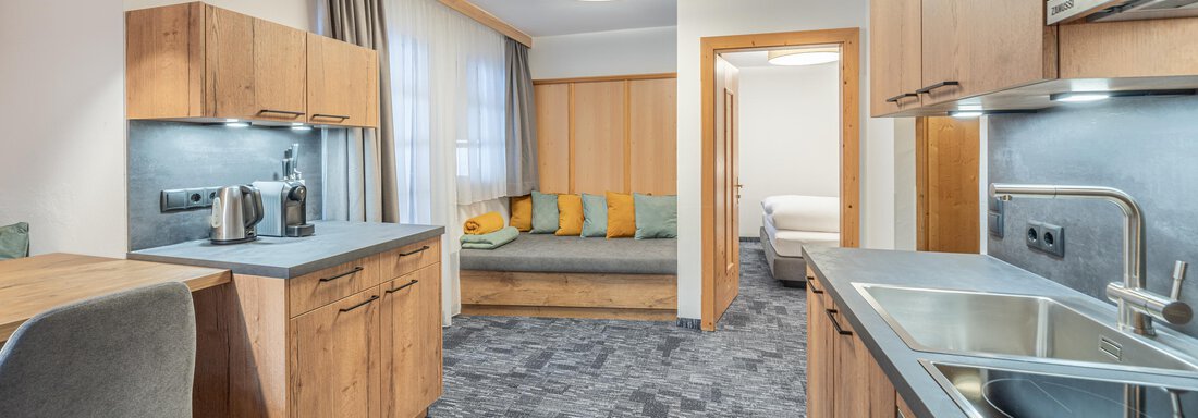 Appartement 17 1-2 Schlafzimmer A modern kitchen with wooden furniture and a cozy living area. The room is bright and inviting, with a comfortable sofa and a large window. - Apart Hotel Garni Alt Kaisers - Sölden
