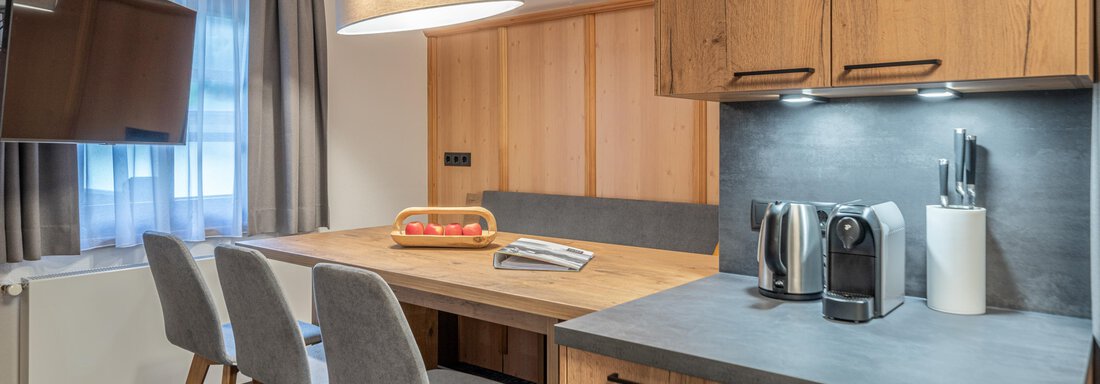 A modern kitchen with wooden cabinets and a dining table. A fruit bowl is on the table and kitchen appliances are visible. - Apart Hotel Garni Alt Kaisers - Sölden