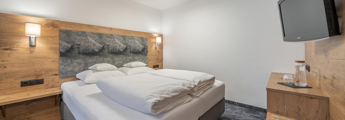 A modern bedroom with a large box spring bed and wooden details. There is a TV on the wall, and the lighting is pleasant. - Apart Hotel Garni Alt Kaisers - Sölden
