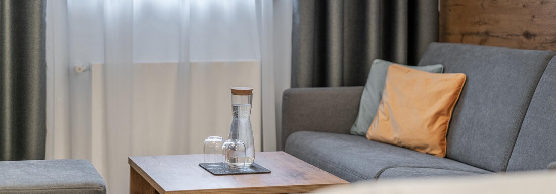A cozy bedroom with a gray sofa and a wooden table. Bright curtains hang in front of the window, and some orange cushions decorate the sofa. - Apart Hotel Garni Alt Kaisers - Sölden