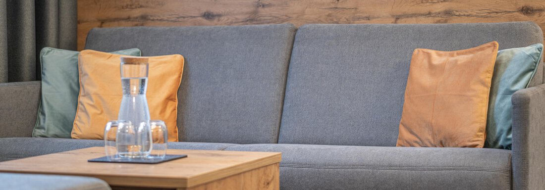 A modern bedroom with a gray sofa and cushions in orange and green. On the table is a glass carafe and a glass. - Apart Hotel Garni Alt Kaisers - Sölden