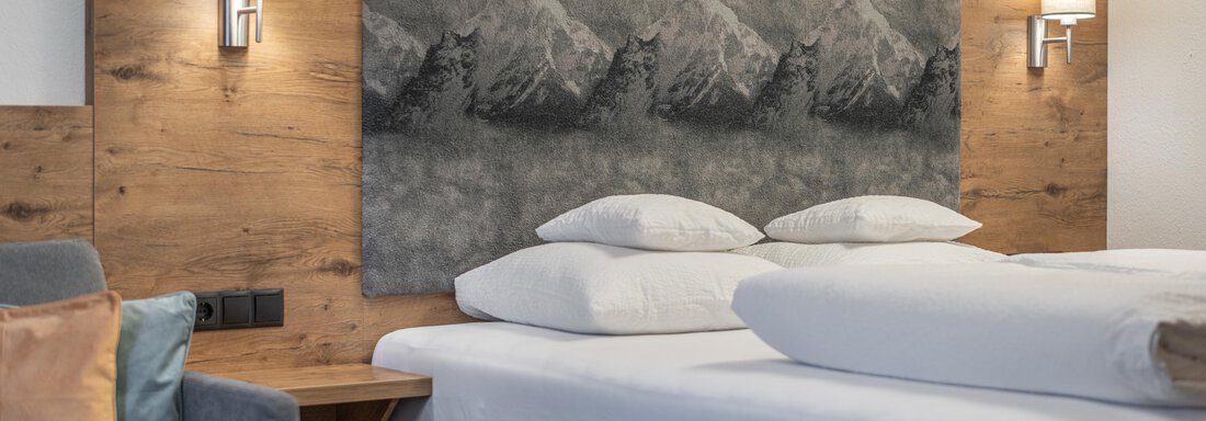 A modern bedroom with a large box spring bed and light cushions. Behind the bed is a decorative wall panel featuring a mountain motif. - Apart Hotel Garni Alt Kaisers - Sölden