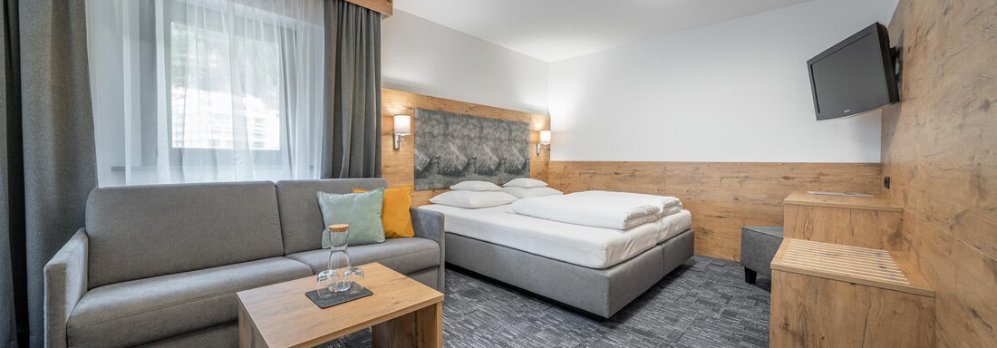 Standard double room | © Johannes Brunner (CC C) A modern hotel room with a box spring bed, a couch, and a wooden table. The walls are light and the carpet is gray, with a large window that lets in natural light. - Apart Hotel Garni Alt Kaisers - Sölden