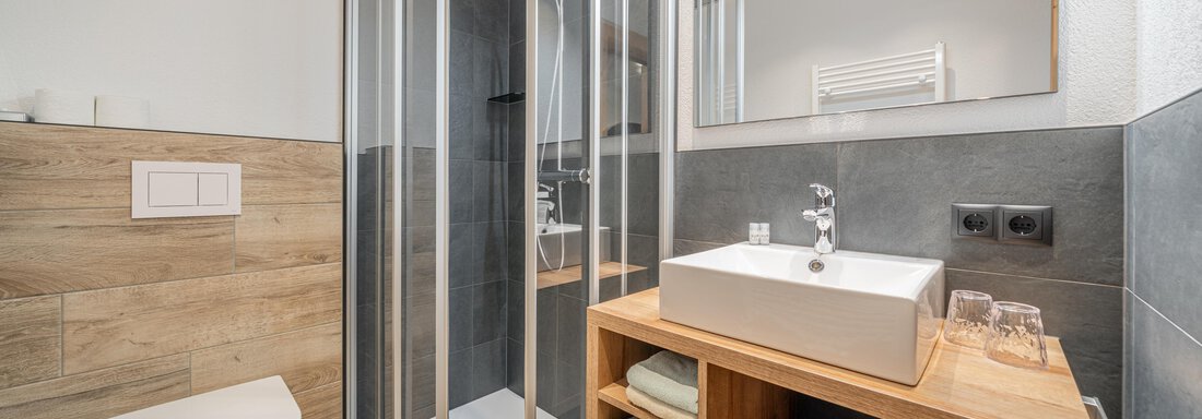 A modern bathroom with a glass shower and an elegant wooden sink unit. The walls are decorated with simple tiles, giving the space a contemporary look. - Apart Hotel Garni Alt Kaisers - Sölden