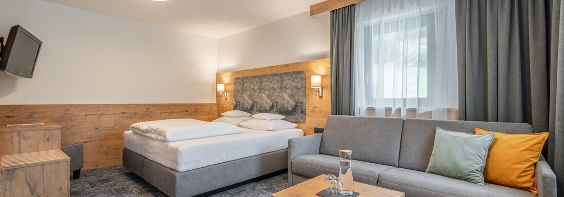 Doppelzimmer Deluxe A modern hotel room with a box spring bed, a sofa, and a wooden table.
Large windows provide plenty of daylight and a pleasant cozy atmosphere. - Apart Hotel Garni Alt Kaisers - Sölden