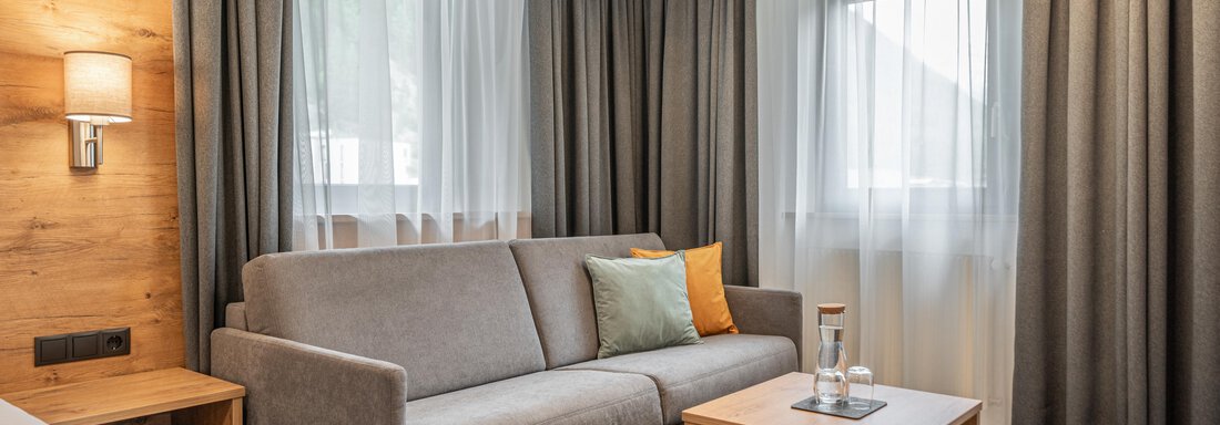 A modern bedroom with a gray sofa and a wooden table. Large windows with curtains provide plenty of daylight. - Apart Hotel Garni Alt Kaisers - Sölden