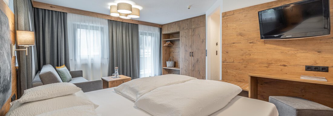 A modern hotel room with a large box spring bed and a cozy seating area. There is a television on the wall and plenty of daylight coming through the windows. - Apart Hotel Garni Alt Kaisers - Sölden