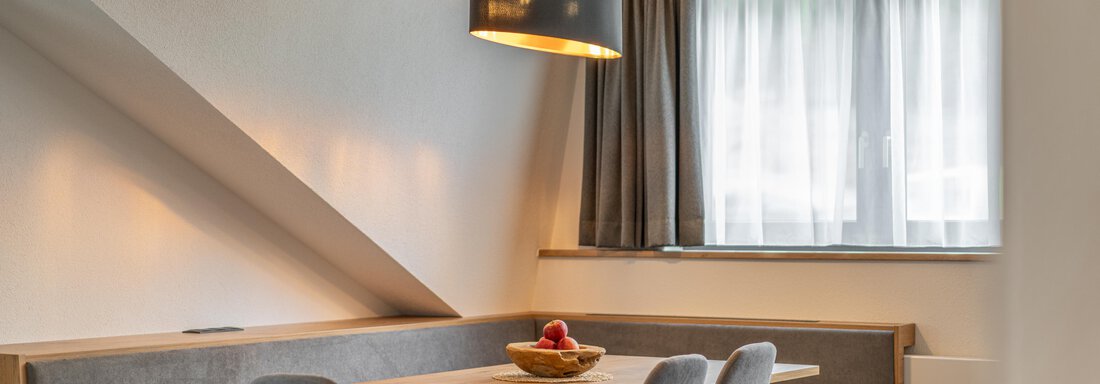 A modern dining room with a wooden slat table and gray chairs. A fresh fruit basket sits on the table, and large windows let in plenty of light. - Apart Hotel Garni Alt Kaisers - Sölden