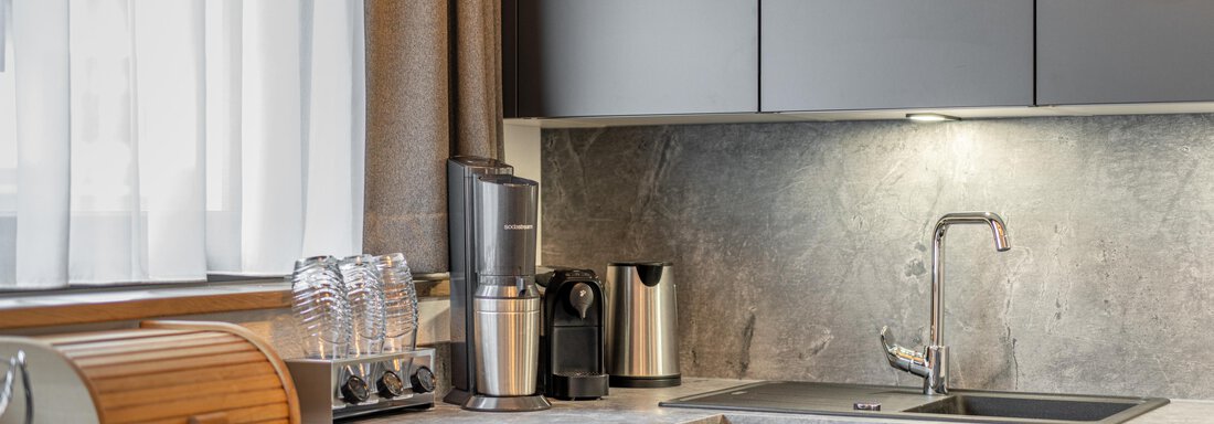 A modern kitchen with gray furniture and a bright countertop. It is well-equipped with appliances and a window that lets in daylight. - Apart Hotel Garni Alt Kaisers - Sölden