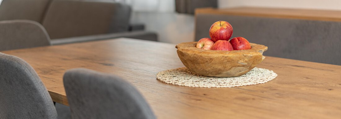 A wooden dining table with a fruit bowl full of apples. In the background, gray chairs and windows with curtains can be seen. - Apart Hotel Garni Alt Kaisers - Sölden