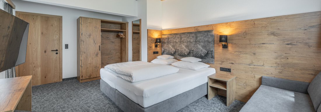 A modern hotel room with a double bed and a gray sofa. The walls are lined with wood, and the room has a bright, inviting atmosphere. - Apart Hotel Garni Alt Kaisers - Sölden