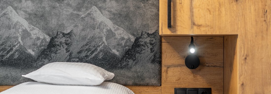 A modern bedroom with a wooden headboard and a stylish nightstand. The wall features a mountain motif and there are cozy pillows placed on the bed. - Apart Hotel Garni Alt Kaisers - Sölden