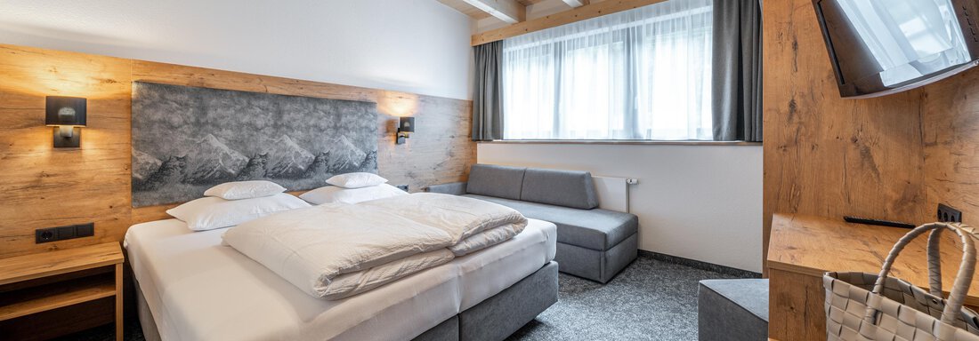 A modern bedroom with a double bed and a seating area. The walls are made of wood and large windows provide plenty of light. - Apart Hotel Garni Alt Kaisers - Sölden