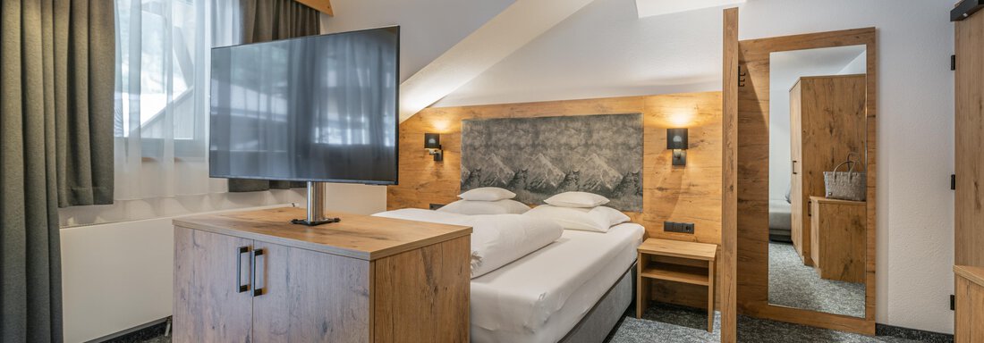 A modern bedroom with a cozy bed and a flat-screen TV. The wooden details and the large window front create a pleasant atmosphere. - Apart Hotel Garni Alt Kaisers - Sölden