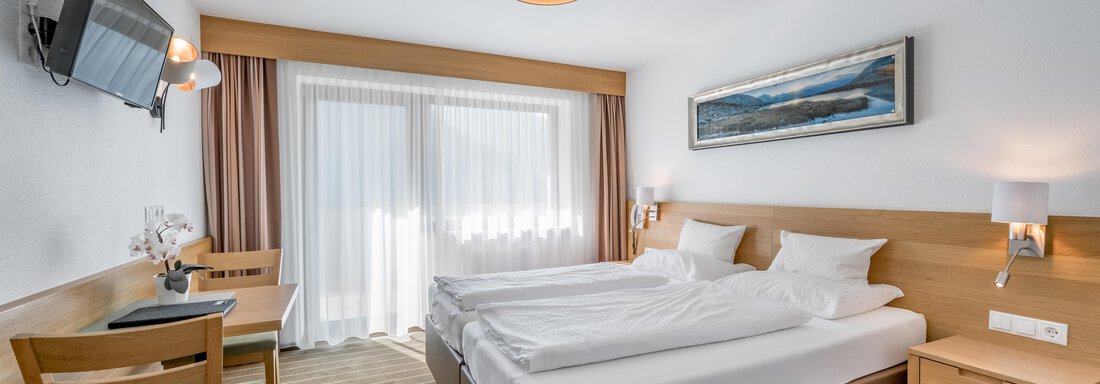 A modern hotel room with two beds, a television, and a table. Large windows provide plenty of natural light and a view of the outdoor area. - Apart Hotel Garni Wieser - Sölden