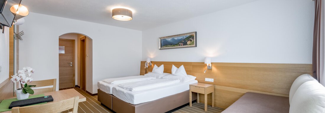 A bright, modern hotel room with two beds and a comfortable sofa. There is a table and a television on the wall. - Apart Hotel Garni Wieser - Sölden